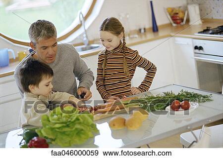 Stock Photo - Father with children in kitchen, helping young son cut vegetables. Fotosearch