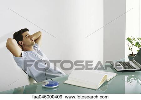 Man at desk leaning back with hands behind head, listening to earphones View Large Photo Image Stock Photo - Man at desk leaning back with hands behind head, listening to earphones. Fotosearch