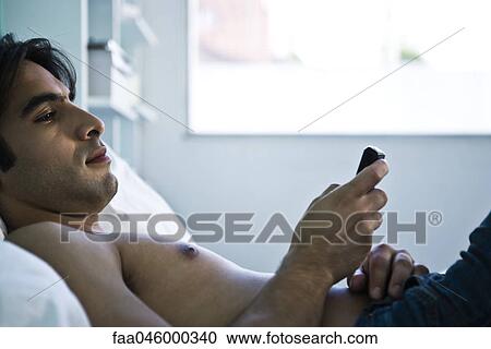 Stock Image - Man reclining on bed looking at cell phone. Fotosearch