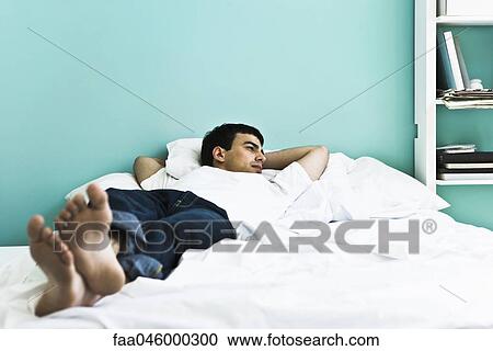 Stock Image - Man reclining on bed with arms behind head. Fotosearch