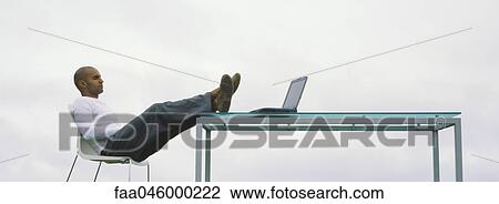 Stock Image - Man sitting outdoors with feet up on table, looking at laptop computer. Fotosearch