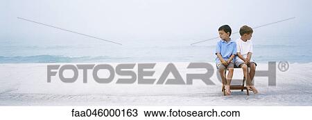 Two boys sitting side by side on folding chair at beach ignoring one another View Large Photo Image Stock Image - Two boys sitting side by side on folding chair at beach ignoring one another. Fotosearch