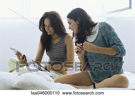 Stock Image - Two female friends sitting in bed looking at magazine together. Fotosearch
