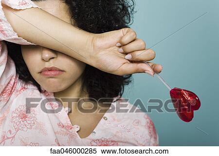 Woman frowning and covering eyes with arm, holding heart-shaped lollipop View Large Photo Image Stock Photography - Woman frowning and covering eyes with arm, holding heart-shaped lollipop. Fotosearch