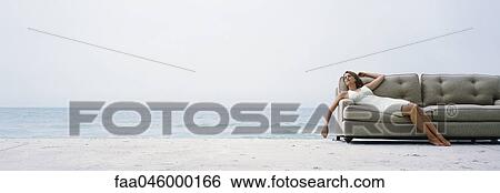 Stock Photograph - Woman reclining on couch on beach at water's edge. Fotosearch