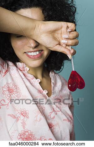Woman smiling, covering eyes with arm, holding heart-shaped lollipop View Large Photo Image Picture - Woman smiling, covering eyes with arm, holding heart-shaped lollipop. Fotosearch