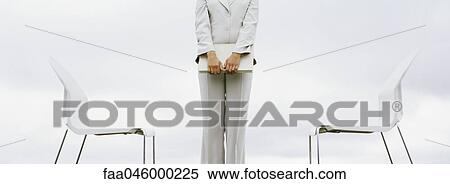 Stock Photography - Woman standing between two chairs holding laptop computer, cropped. Fotosearch