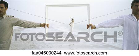 Picture - Woman walking on beach, framed by picture frame held aloft by two men in foreground. Fotosearch