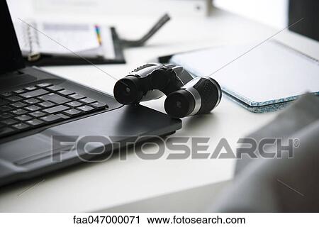 Stock Image - Binoculars on desk with laptop computer and notebook. Fotosearch