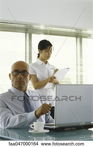 Picture - Businessman sitting at desk, looking at camera, female assistant taking notes in background. Fotosearch
