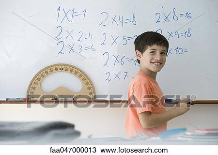 Stock Image - Elementary school student writing equations on whiteboard, smiling over shoulder at camera. Fotosearch