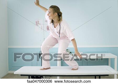 Stock Photograph - Female medical student standing on top of examination table, pretending to surf. Fotosearch