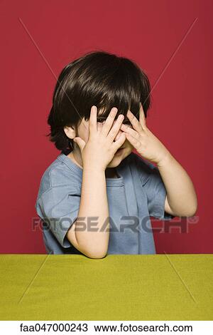 Stock Image - Little boy covering face with hands, peeking through fingers at camera. Fotosearch