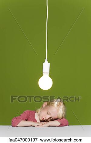 Stock Photo - Little girl asleep resting head on arms, illuminated light bulb suspended overhead. Fotosearch