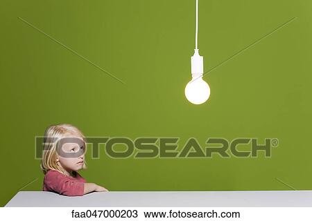 Stock Image - Little girl biting lip looking with concern up at illuminated light bulb suspended overhead. Fotosearch