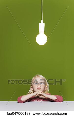 Stock Photo - Little girl contemplating illuminated light bulb suspended overhead. Fotosearch