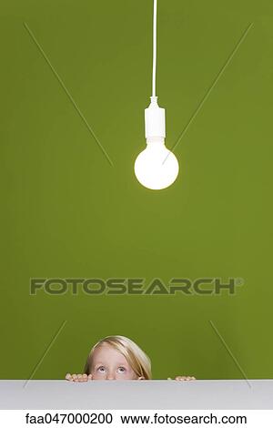 Stock Image - Little girl looking fearfully up at illuminated light bulb suspended overhead. Fotosearch