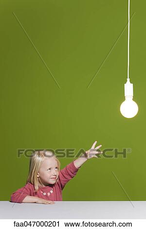 Stock Image - Little girl reaching for illuminated light bulb suspended overhead. Fotosearch