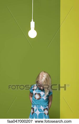 Stock Photo - Little girl with hands behind back contemplatively looking down, illuminated light bulb suspended overhead. Fotosearch