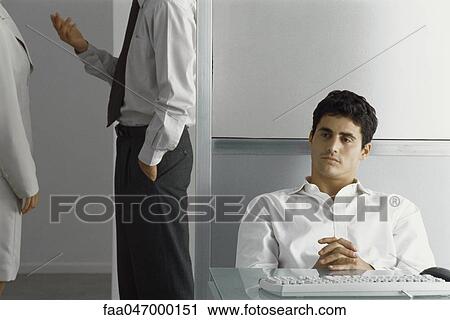 Man sitting at desk with hands clasped while colleagues talk nearby View Large Photo Image Stock Image - Man sitting at desk with hands clasped while colleagues talk nearby. Fotosearch