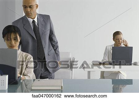 Stock Image - Manager standing, looking over shoulder of female employee while she works. Fotosearch