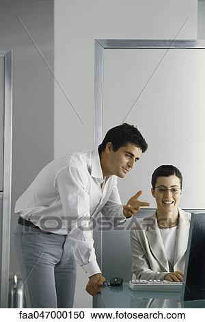 Stock Image - Office scene, man leaning on edge of desk speaking to female colleague, both looking at her computer screen. Fotosearch
