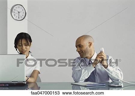 Office worker sitting at desk holding phone, covering receiver and speaking to female co-worker View Large Photo Image Stock Image - Office worker sitting at desk holding phone, covering receiver and speaking to female co-worker. Fotosearch