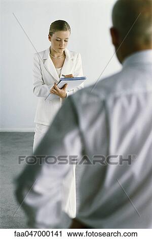 Stock Image - Young assistant standing taking notes, boss dictating in foreground. Fotosearch