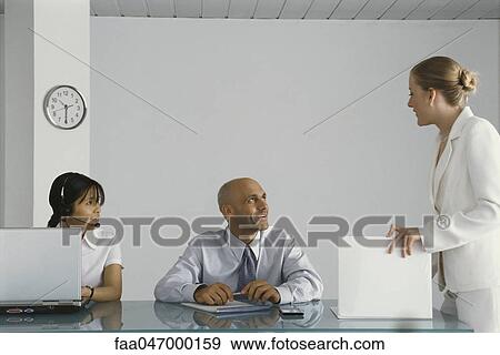 Stock Photo - Young female executive standing by colleagues' desk, conversing. Fotosearch