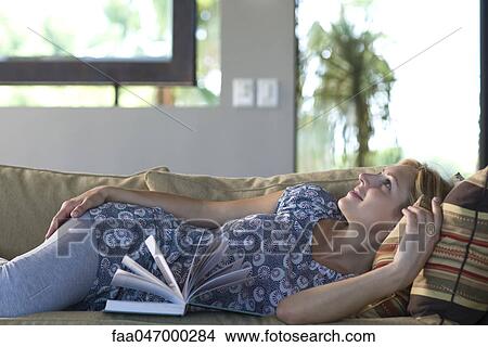 Picture - Young woman reclining on sofa, dreamily looking away with book at side. Fotosearch