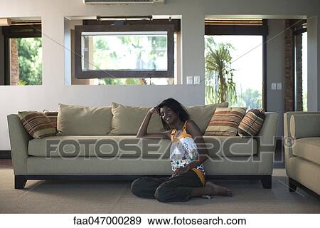 Stock Photo - Young woman sitting on living room floor leaning against sofa, smiling. Fotosearch