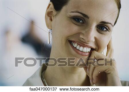 Stock Photo - Young woman smiling at camera, leaning face against hand, portrait. Fotosearch