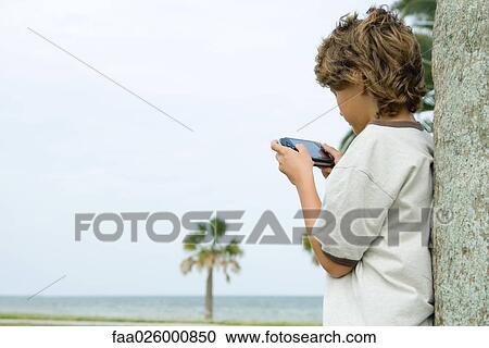 Boy leaning against tree trunk, playing handheld video game, side view View Large Photo Image Stock Image - Boy leaning against tree trunk, playing handheld video game, side view. Fotosearch