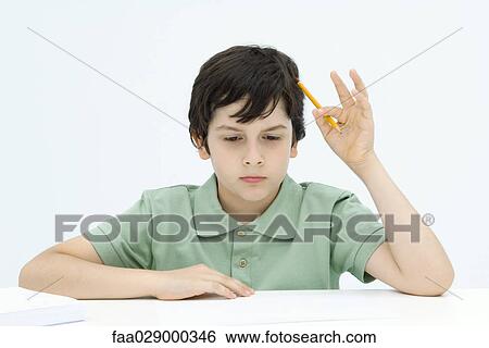 Stock Photograph - Boy sitting at table, holding pencil against head, looking down. Fotosearch