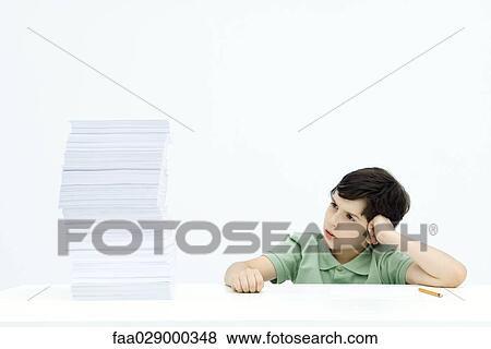 Stock Photo - Boy sitting at table next to tall stack of homework, frowning. Fotosearch