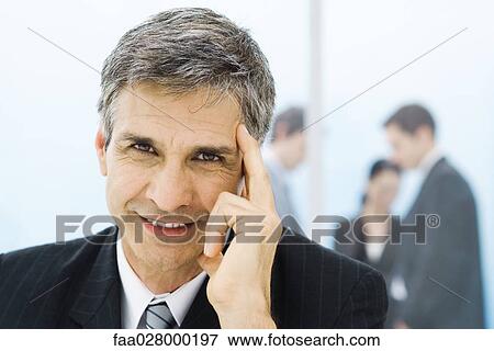 Businessman holding head, smiling at camera, colleagues in background View Large Photo Image Stock Photo - Businessman holding head, smiling at camera, colleagues in background. Fotosearch