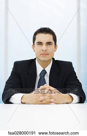 Businessman sitting at table with clasped hands, looking at camera View Large Photo Image Stock Image - Businessman sitting at table with clasped hands, looking at camera. Fotosearch