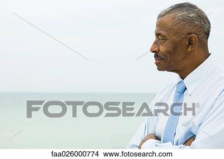 Picture - Businessman smiling with arms folded, side view, sea in background. Fotosearch