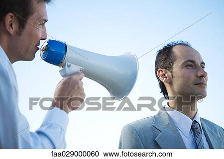Stock Image - Businessman standing beside colleague, shouting into megaphone. Fotosearch
