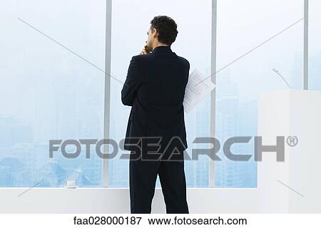 Businessman standing in front of window, looking out, holding document, rear view View Large Photo Image Stock Photo - Businessman standing in front of window, looking out, holding document, rear view. Fotosearch