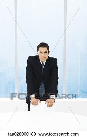 Stock Photo - Businessman standing with hands on chair, looking at camera. Fotosearch