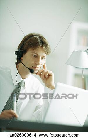 Businessman wearing headset, looking at laptop computer, hand under chin View Large Photo Image Picture - Businessman wearing headset, looking at laptop computer, hand under chin. Fotosearch