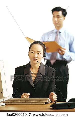 Stock Photo - Businesswoman sitting at desk, wearing headset, looking away. Fotosearch