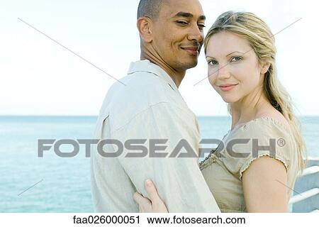 Couple embracing, both smiling at camera, ocean in background View Large Photo Image Stock Image - Couple embracing, both smiling at camera, ocean in background. Fotosearch