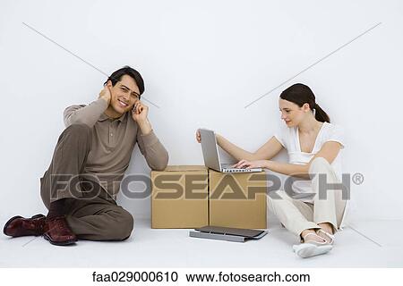 Stock Image - Couple sitting at makeshift desk, woman using laptop computer, man using cell phone and smiling at camera. Fotosearch