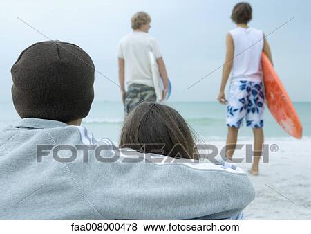 Couple sitting on beach looking toward two surfers looking at waves, rear view View Large Photo Image Stock Photo - Couple sitting on beach looking toward two surfers looking at waves, rear view. Fotosearch