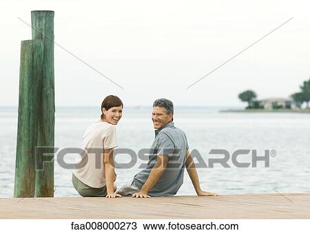 Couple sitting on edge of dock, smiling over shoulders at camera View Large Photo Image Stock Image - Couple sitting on edge of dock, smiling over shoulders at camera. Fotosearch