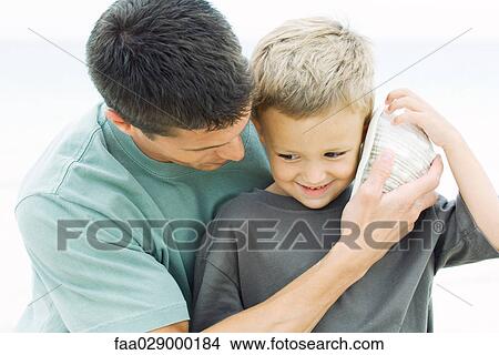 Father holding seashell up to young son's ear at the beach, close-up View Large Photo Image Picture - Father holding seashell up to young son's ear at the beach, close-up. Fotosearch