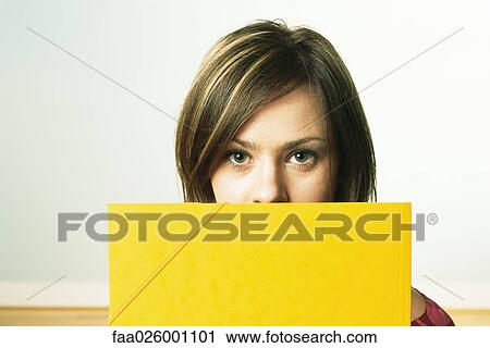Stock Image - Female holding folder in front of face, looking at camera. Fotosearch
