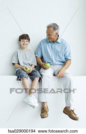 Grandfather and grandson sitting side by side on ledge, holding apples, both smiling View Large Photo Image Picture - Grandfather and grandson sitting side by side on ledge, holding apples, both smiling. Fotosearch